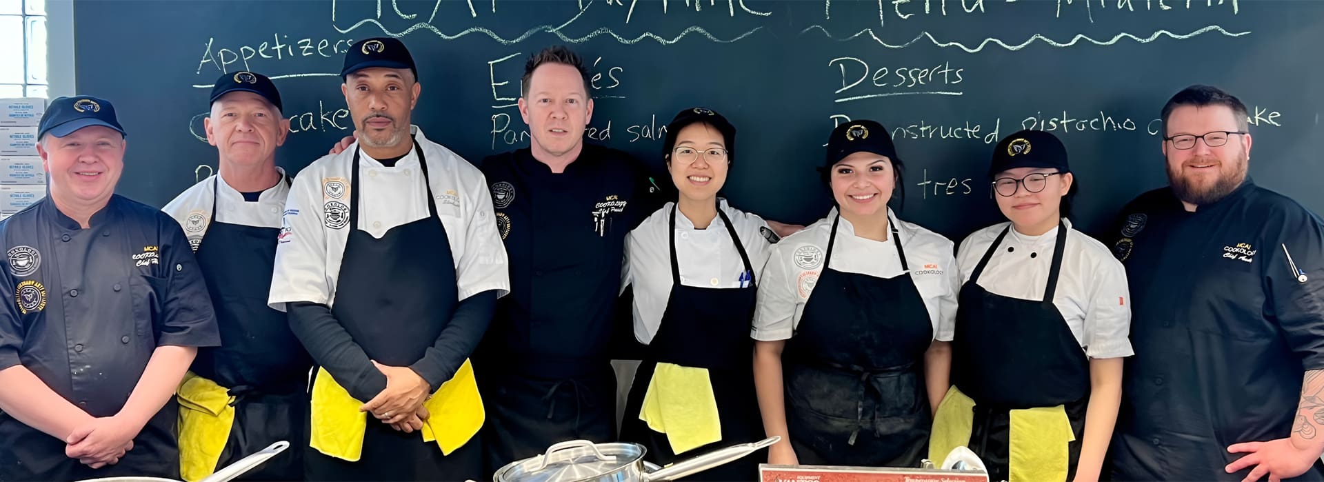 A group of chefs in uniforms stands in front of a chalkboard menu.