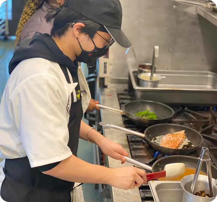 Person wearing a black cap and mask cooks salmon in a pan on a stove in a kitchen. Another person in the background prepares food.