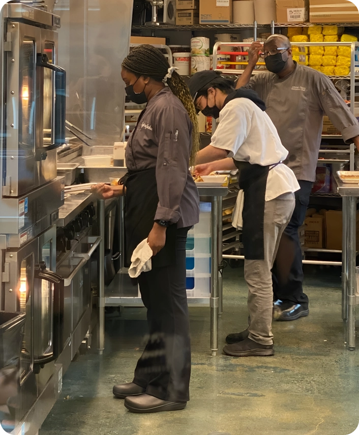 Three chefs in a kitchen, wearing uniforms and masks, focus on preparing food. Stainless steel appliances and shelves stocked with ingredients are visible.