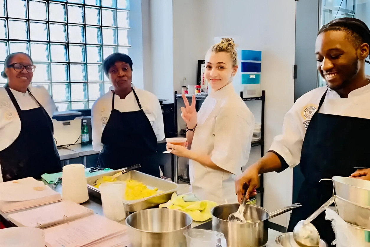 Four chefs in a kitchen, with one giving a peace sign, preparing food at a counter.